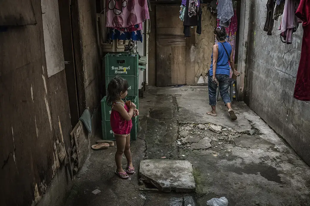 Josie, a 43-year-old former "shabu" (methamphetamine) user and pusher, navigates the living area of the bar in a rural area of the Philippines, where she works as a housekeeper. She is being watched by one of the bar worker's daughters. Recently Josie fled the Market 3 slum in Navotas because she feared assassins might come to execute her. Her drug-related activities were well known in Market 3 and many others have been executed for similar activities. Image by James Whitlow Delano. Philippines, 2017.