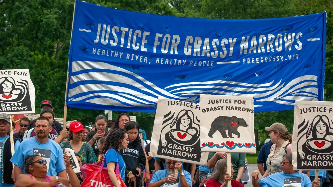 A group of Grassy Narrows First Nation residents and their supporters stand below a "Justice for Grassy Narrows" banner. Members of the Grassy Narrows First Nation traveled to Toronto to demand justice. Image by Shelby Gilson. Canada, 2019. A group of Grassy Narrows First Nation residents and their supporters stand below a "Justice for Grassy Narrows" banner. Members of the Grassy Narrows First Nation traveled to Toronto to demand justice. Image by Shelby Gilson. Canada, 2019.