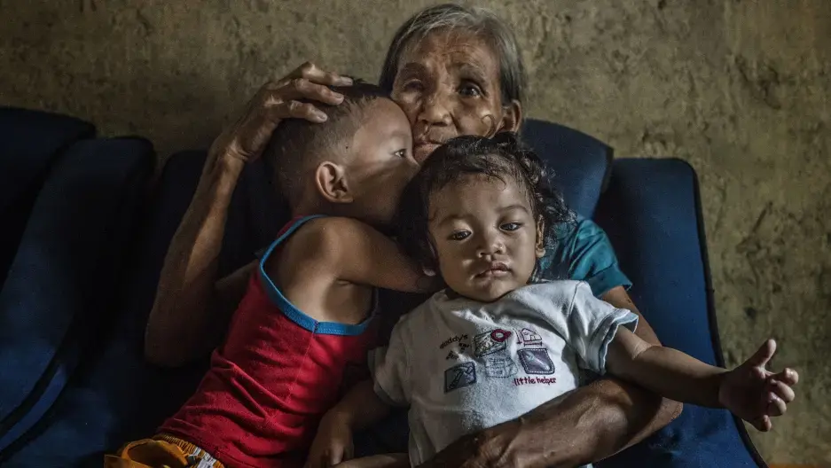 Remy Fernandez, 84 years old, holds two of the seven grandchildren she is raising. Her youngest son, Constantino de Juan, a methamphetamine user, was killed by masked men in December, 2016. Upon seeing his attackers, Juan instructed five-year-old CJ, shown here wearing a red tank top, to take care of his siblings. The children's mother is in prison due to a drug arrest. Baby RJ, in the "daddy's little helper" T-shirt, was born in prison. Image by James Whitlow Delano. Philippines, 2017.