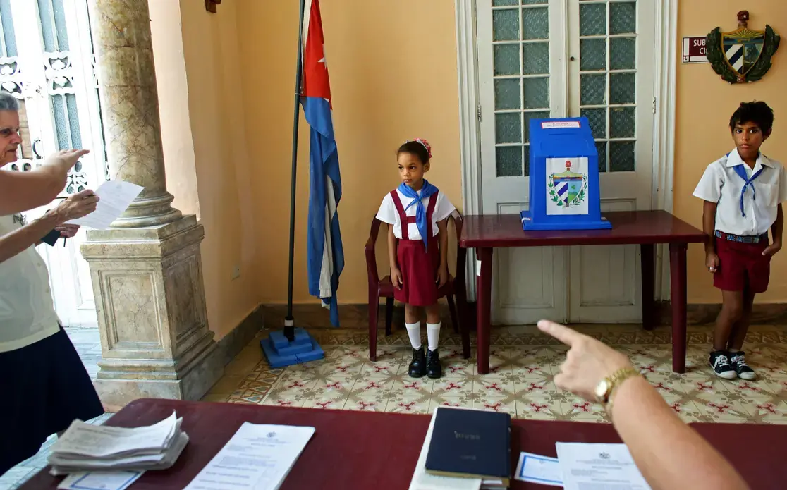 Children wearing the uniform of Communist youth are prepared to salute “Votó!” (“He voted!”), as a woman places her ballot in Cuba’s Elecciones Parciales (Partial Elections). The vote was to elect delegates to the Municipal Assemblies of People’s Power, the country’s unicameral parliament, on April 19, in Havana. The delegates function as district representatives for a two-and-a-half-year term. Image by Yana Paskova. Cuba, 2015. Children wearing the uniform of Communist youth are prepared to salute “Votó!” (“He voted!”), as a woman places her ballot in Cuba’s Elecciones Parciales (Partial Elections). The vote was to elect delegates to the Municipal Assemblies of People’s Power, the country’s unicameral parliament, on April 19, in Havana. The delegates function as district representatives for a two-and-a-half-year term. Image by Yana Paskova. Cuba, 2015.