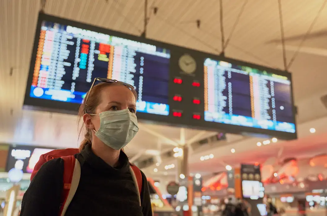 A woman with a backpack wears an anti-virus mask on her face at an airport. Image by Damian Pankowiec/Shutterstock. 2020. A woman with a backpack wearing an anti-virus mask on her face at the airport arrivals board. Image by Damian Pankowiec/Shutterstock. 2020.
