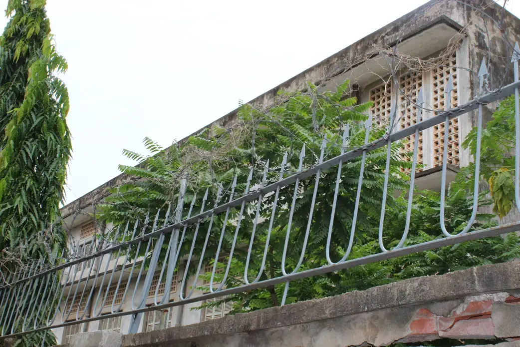 Perimeter of S-21 in Phnom Penh, the former school that was converted into a concentration camp during the Khmer Rouge era. Image by M.G. Zimeta. Cambodia, 2018. Photograph of S-21 former concentration camp, taken from the perimeter wall. Barbed wires, spiked metal railings, and overgrown ferns partly shield the view of the building. Image by M.G. Zimeta. Cambodia, 2018.
