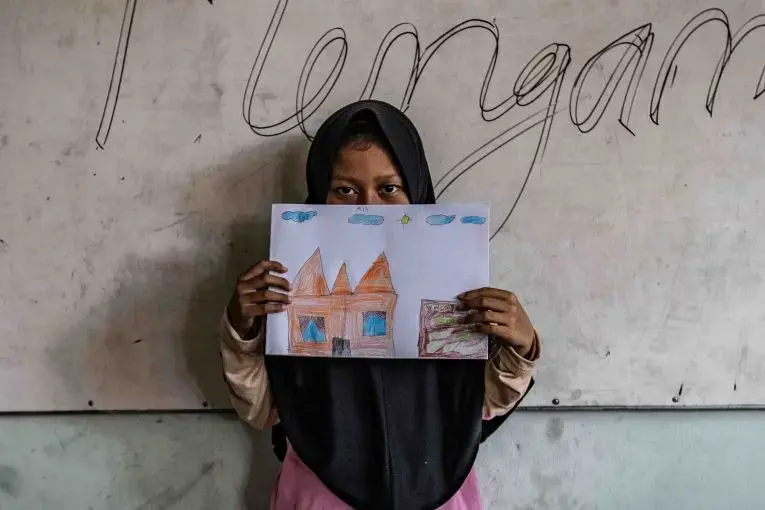A 10-year old girl holds up her drawing of a home in a government run shelter. The girl is one of the surviving members of the families that carried out a string of ISIS-inspired suicide attacks in the city of Surabaya in May 2018. Image by Jurnasyanto Sukarno. Indonesia, 2018. A 10-year old girl holds up her drawing of a home in a government run shelter. The girl is one of the surviving members of the families that carried out a string of ISIS-inspired suicide attacks in the city of Surabaya in May 2018. Image by Jurnasyanto Sukarno. Indonesia, 2018.