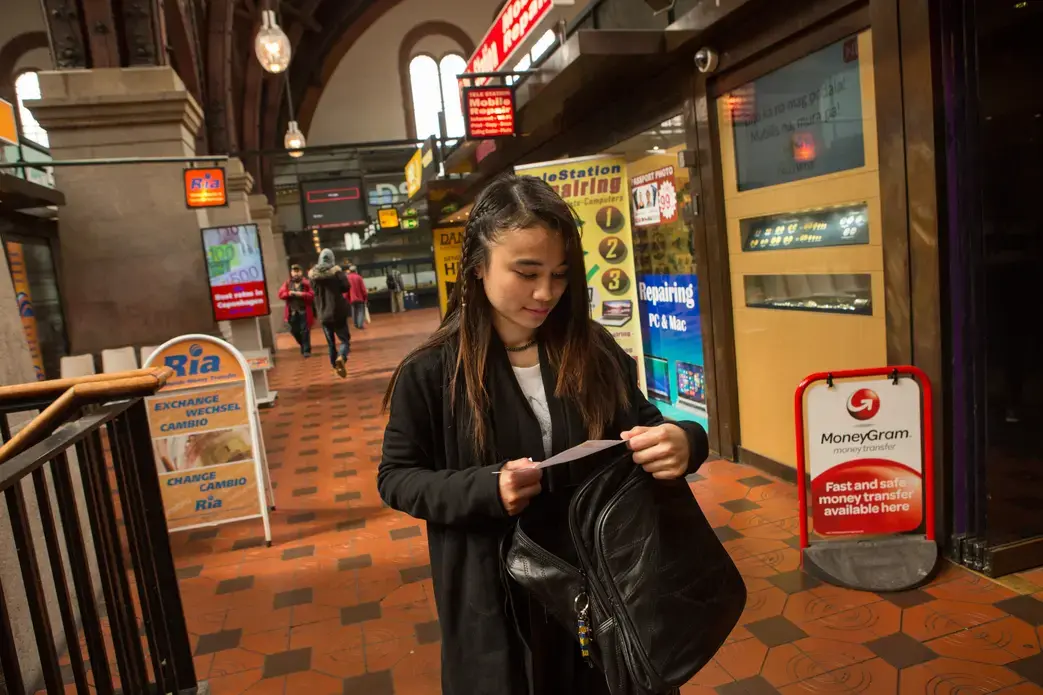 Au pair Lucila Espinosa Milay checks her receipt as she walks through a row of money transfer businesses after wiring part of her paycheck to siblings back in the Philippines. Milay pays the school fees for several of her eight siblings back home, sending money every 15 days. Remittances are a huge economic driver in the Philippines. Image by Allison Shelley. Denmark, 2016. Au pair Lucila Espinosa Milay checks her receipt as she walks through a row of money transfer businesses after wiring part of her paycheck to siblings back in the Philippines, at Central Station in Copenhagen, Denmark, May 21, 2016. Milay pays the school fees for several of her eight siblings back home, sending money every 15 days. Remittances are a huge economic driver in the Philippines. Image by Allison Shelley. Philippines, 2016.