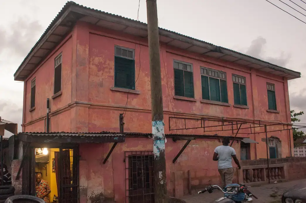 Tailor Pierre Kwetey inside his workshop, as a man walks by the house that allegedly contained a fake US Embassy in Accra. Image by Yepoka Yeebo. Ghana, 2017.