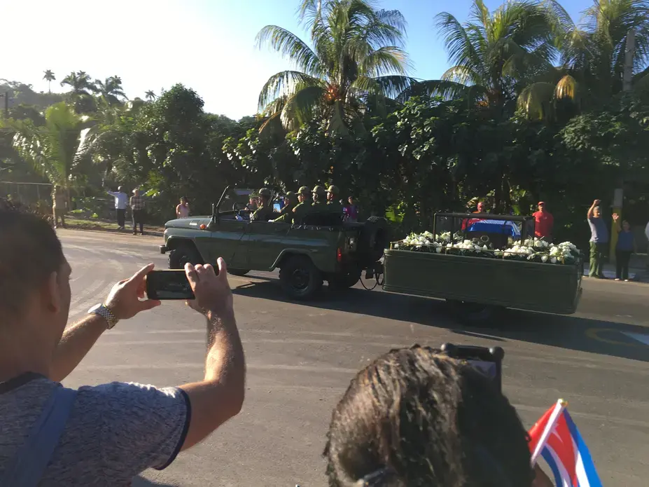 The remains of Fidel Castro traveled along a 500 mile journey from Havana to Santiago, his final resting place—the exact reverse journey Castro took when he launched his 1959 revolution. Image by Zach Fannin. Cuba, 2016. Fidel Castro's remains are transported 500 miles from Havana to Santiago. The image shows an open car pulling a trailer holding his remains, surrounded by flowers.
