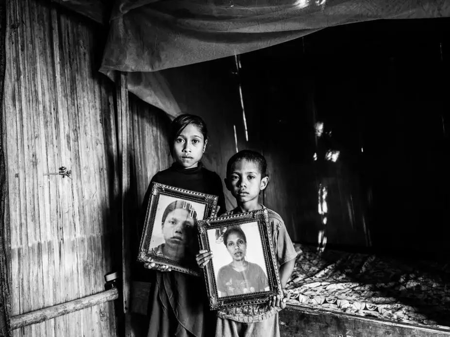 Dolfina’s children Erna and Julius hold photos of their mother inside their hut in East Nusa Tenggara, Indonesia. They now live with their grandfather Mikhael. Image by Xyza Bacani. Indonesia, 2018.