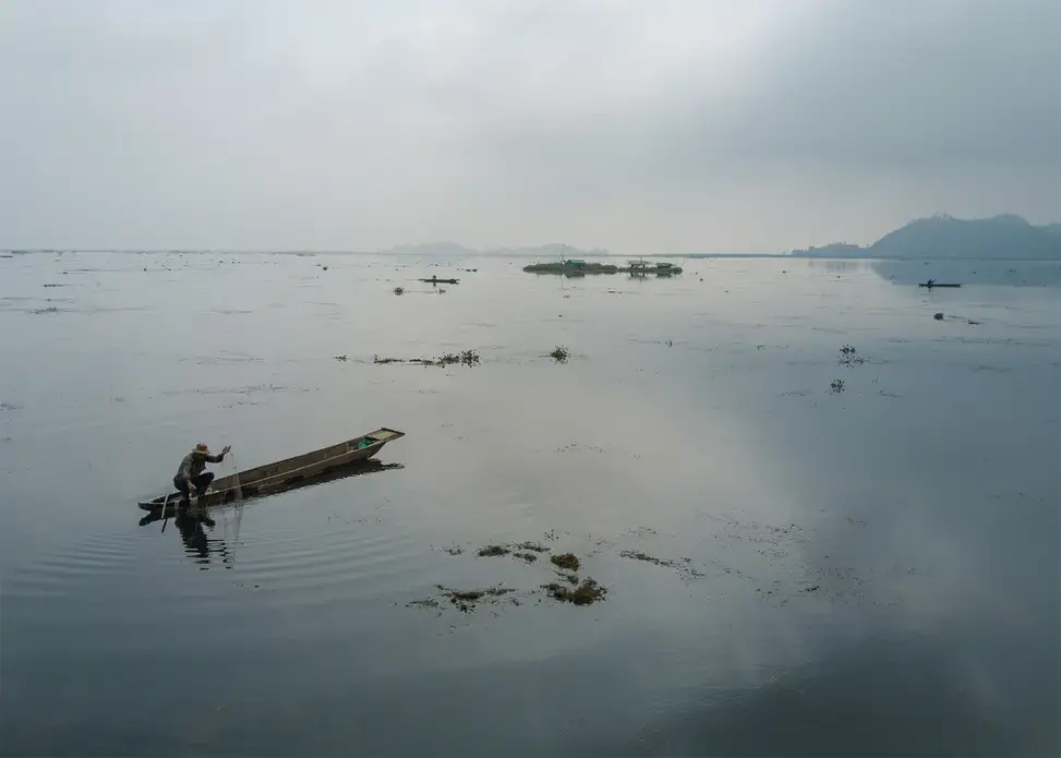 A Meitie fisherman fishes near phumshangs (floating huts) in Loktak Lake, Manipur. The construction of the Ithai Dam flooded over 80,000 hectares of farm and pasture land in the vicinity of the Loktak wetland. The Loktak Development Authority has been dredging the phumdis (floating biomass) and has cleared over 800 Phumshangs in the name of lake clean-up for efficient power generation. Image by Neeta Satam. India, 2017.