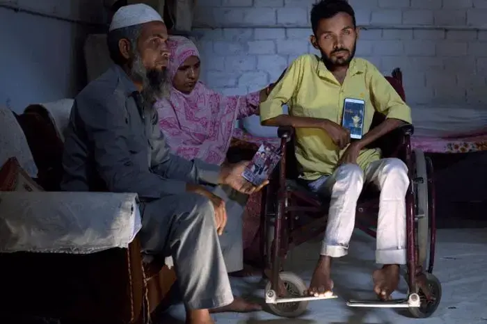 Umar Khan, 24, with his parents shows photos of his younger brother Azhar who died last year. Azhar was suffering from muscular dystrophy as is Umar. Umar watched his younger brother dying just last year and he knows that is coming to him in a few short years. Umar’s parents are very caring towards him. They feed him, wash him and help him in the toilet. He needs 24 hour care. That’s what they were doing for both children until last year. Image by Rohit Jain. India, 2019.