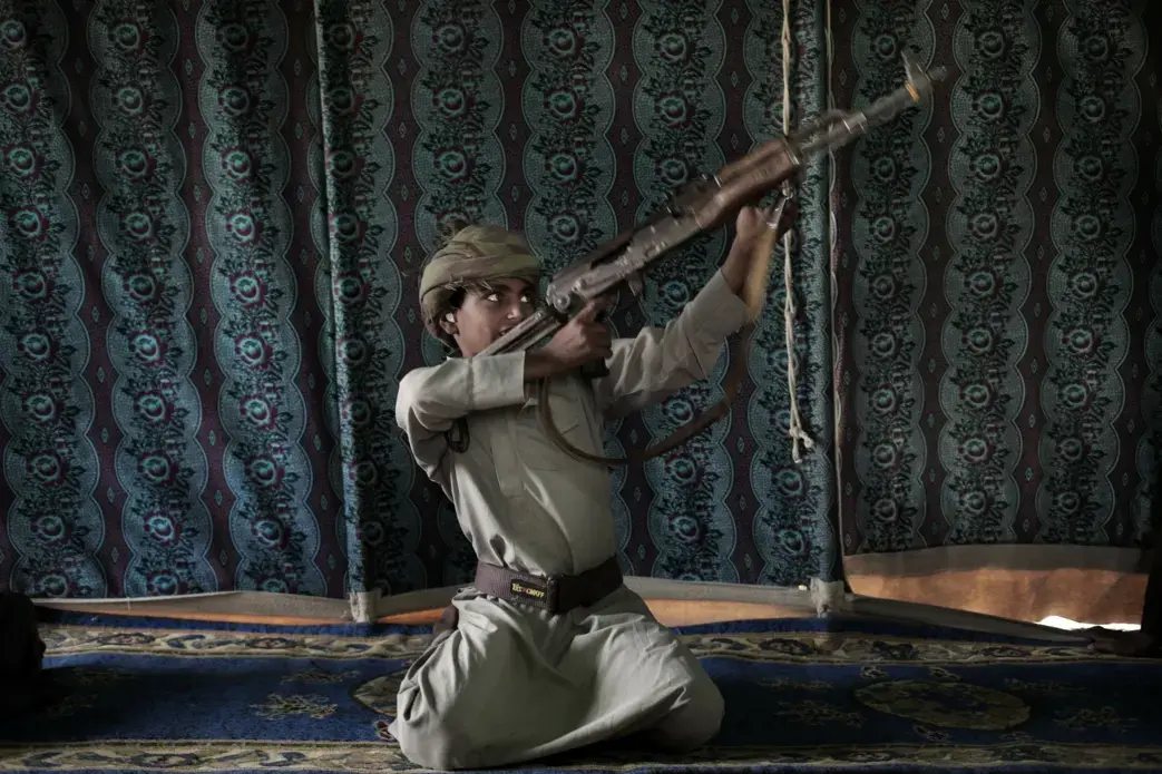 Kahlan, a 12-year-old former child soldier, demonstrates how to use a weapon, at a camp for displaced persons where he took shelter with his family, in Marib, Yemen in this July 27, 2018 photo. Houthi rebels took Kahlan and his classmates, promising to give them new school bags, but instead they were enlisted and trained as fighters tasked with carrying supplies to the front lines. There, he had to elude explosions and airstrikes that left mangled casualties on the battlefield. "The sight of the bodies was scary," he said. Image by Nariman El-Mofty / AP News. Yemen, 2018. Kahlan, a 12-year-old former child soldier, demonstrates how to use a weapon, at a camp for displaced persons where he took shelter with his family, in Marib, Yemen in this July 27, 2018 photo. Houthi rebels took Kahlan and his classmates, promising to give them new school bags, but instead they were enlisted and trained as fighters tasked with carrying supplies to the front lines. There, he had to elude explosions and airstrikes that left mangled casualties on the battlefield. "The sight of the bodies was…