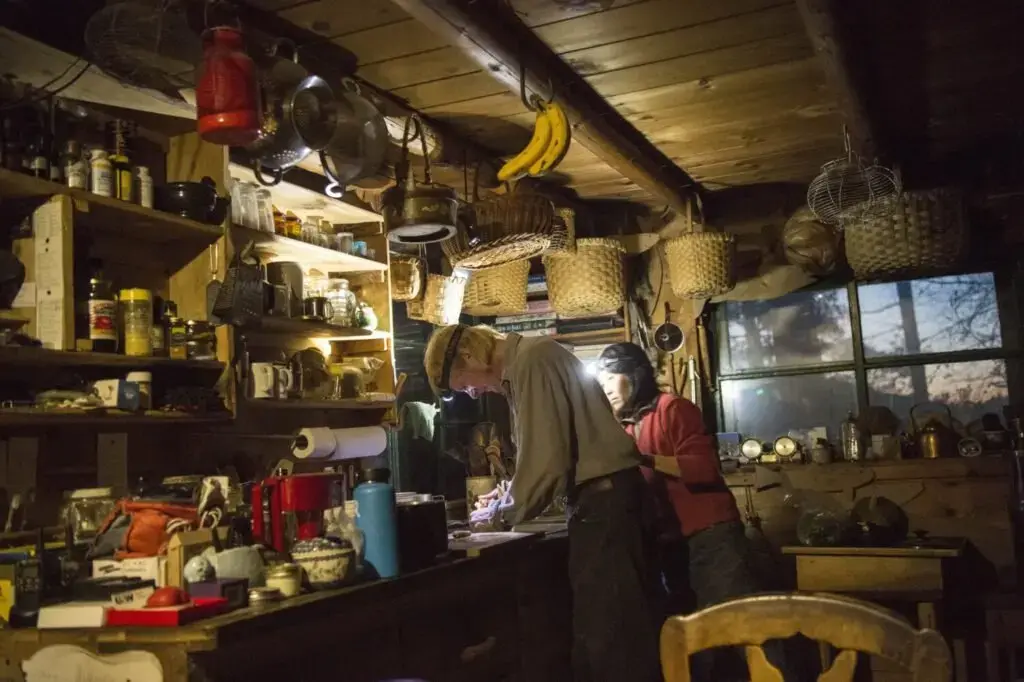 Duane Hanson and Sally Kwan prepare dinner by headlamp at their home on Whipple Pond in T5 R7 in the Unorganized Territories of Maine on May 26, 2019. The homestead is equipped with one solar panel that powers a satellite phone charger and one light bulb over the kitchen table. Image by Michael G. Seamans. United States, 2019.