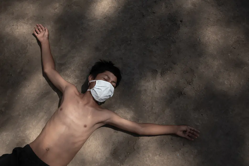 A young boy lies down on the dirt ground with arms outstretched. He is wearing a white surgical mask, black shorts, and no shirt.