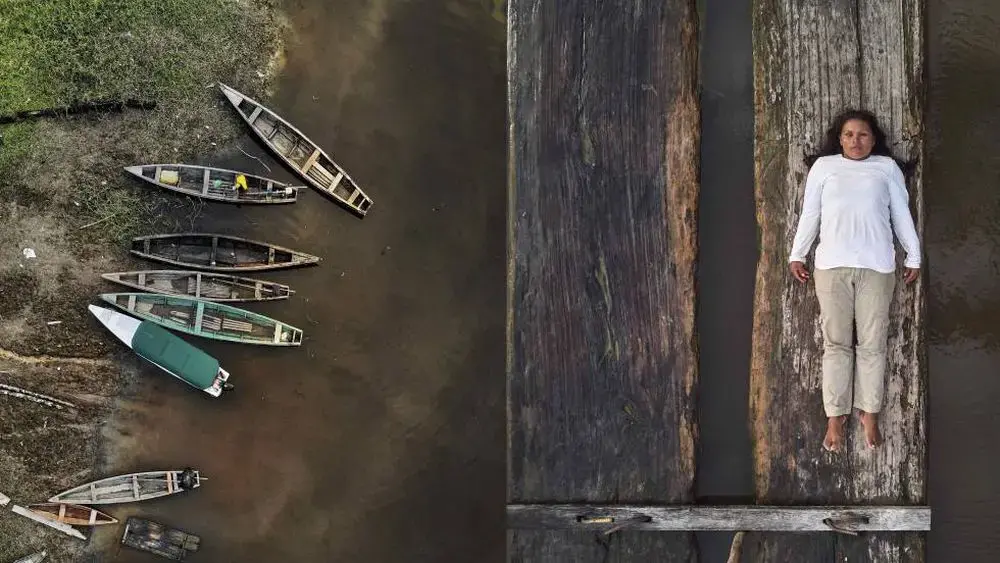 To the left, a series of empty wooden canoes stand by the margins of a river. On the right, Lilia Isolina Java Tapayuri lies down on a plank of wood, looking directly at the camera, captured from an aerial shot. She is wearing a white long sleeved shirt, khaki-colored pants, and no shoes.