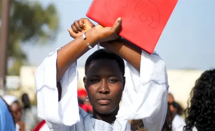 Image by Ricci Shryock. Senegal, 2012. Student dressed in white makes an X with arms above head while holding a red folder
