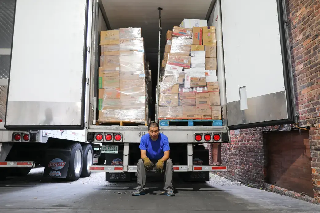 A truck driver leans on the edge of his 18-wheeler, which is full of boxes.