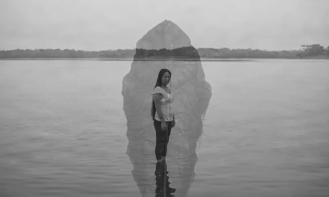 Imagem de Florence Goupil. Black and white double exposure photo of a woman standing in front of a body of water with a leaf superimposed on the image.