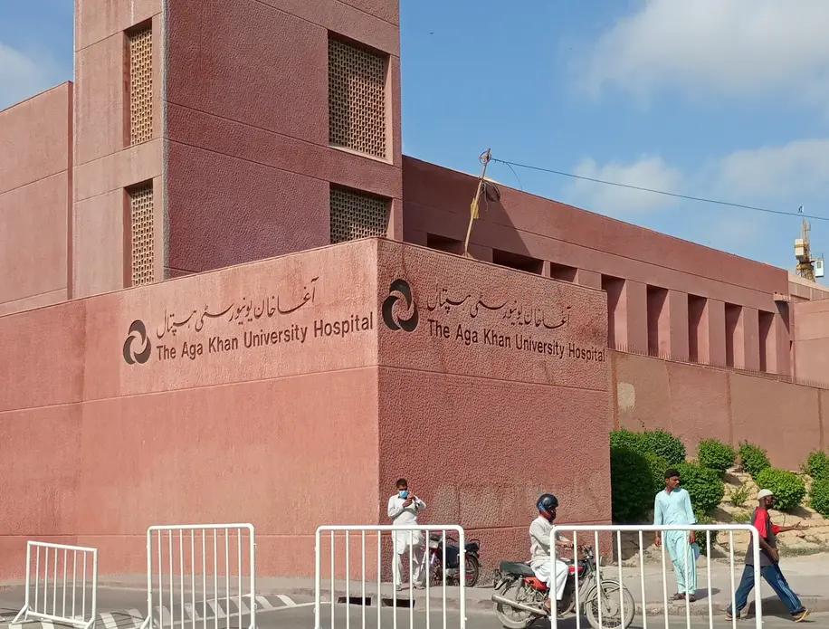 Image of people in front of hospital building. On the building, "Aga Khan University Hospital" is written in English and Urdu.