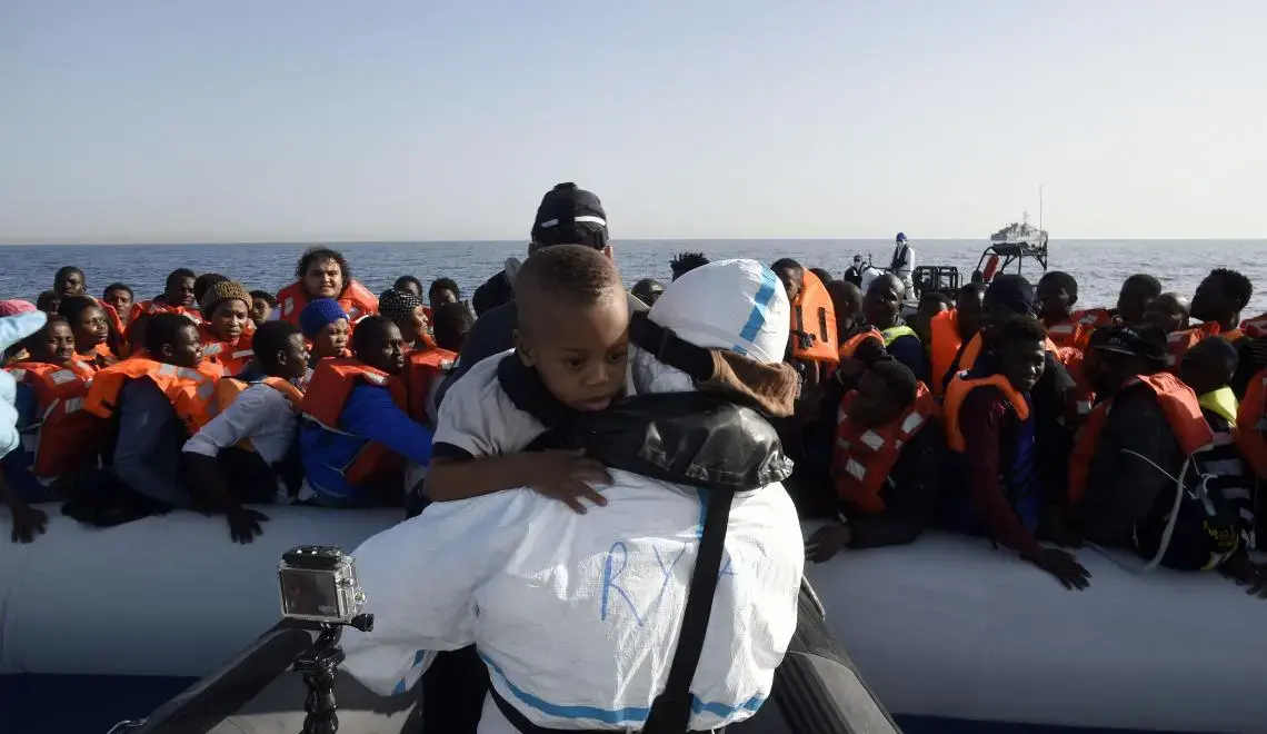 Dozens of Africans are crammed in a raft, all wearing life vests. Beyond them is the sea. In the foreground in front of the raft, someone wearing a white, waterproof uniform is carrying a little boy who was presumably on the raft. The boy looks over the person's shoulder, toward the camera.