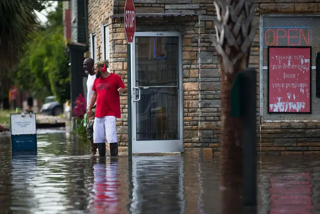 Two men stand outside a store by a stop sign with floodwaters reaching past their ankles. One man is wearing a red t-shirt, white shorts, and knee-high rubber boots. The other man is wearing a white t-shirt and pants.