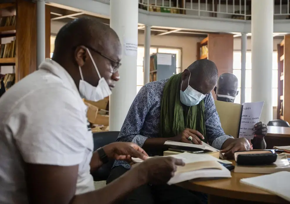Three people wearing medical face masks sit and read papers and books in an archive.