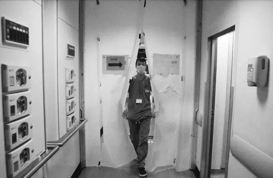 Black and white photo of a man wearing scrubs and a white face mask walking through white curtains that have a sign on them saying "keep out."