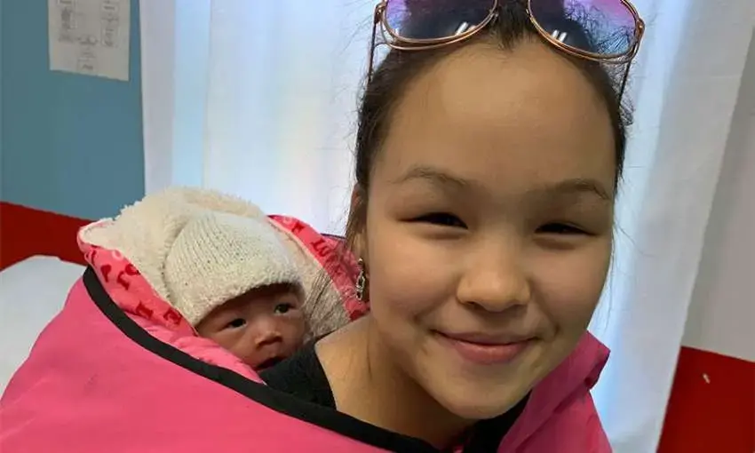 Image of a smiling young woman with an infant secured on her back with a pink blanket. The woman has sunglasses propped on her head, and the infant is wearing a white knit hat.