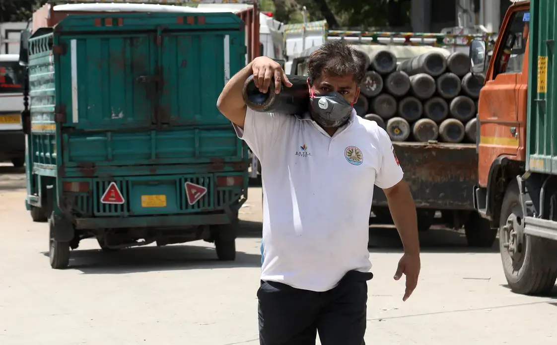 A man carries an oxygen cylinder. There are trucks in the background.