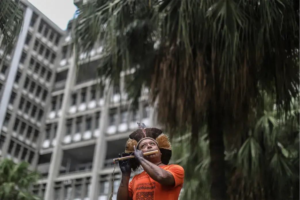 A man in traditional Indigenous dress stands in front of a tall building and plays a flute-like instrument.