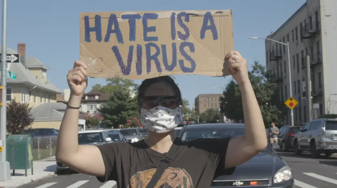 Film still 2.png A masked person holds a sign that reads 'hate is a virus'