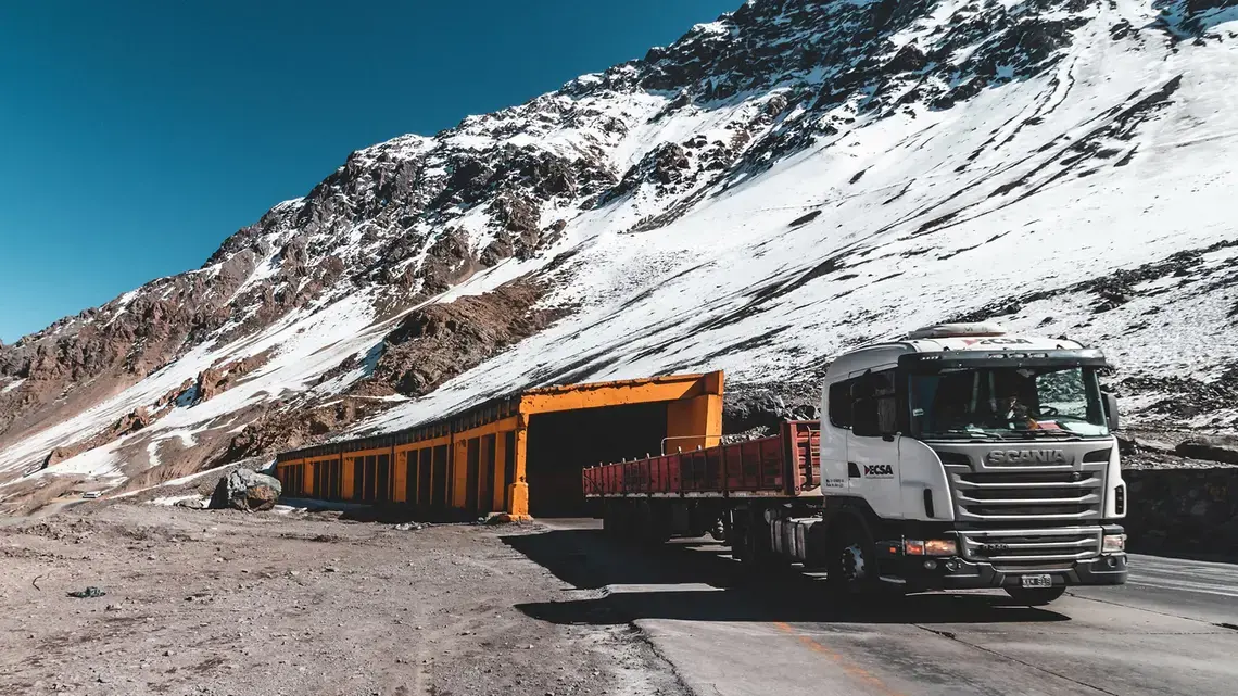 A large white truck drives through a yellow tunnel at the base of snow-dusted mountains.