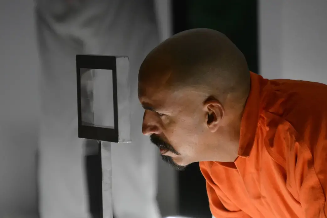 Actor in an orange jumpsuit looks out meal slot set piece.