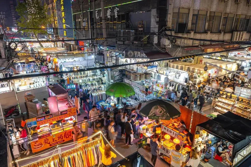 stalls are set up in rows outdoors in an urban area at night. Different colored lights make the squares of stalls glow in tones of red, yellow, and green.