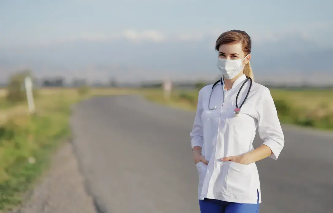 a woman wearing a white medical coat and a face mask stands on an empty road with flat green expanses behind her