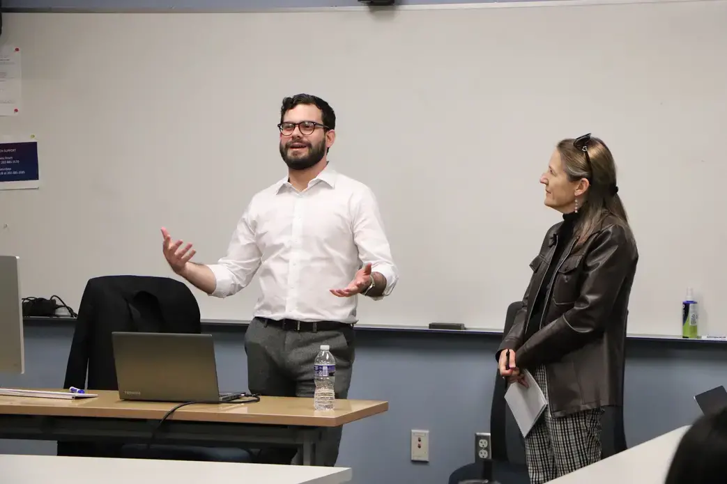 Man in white shirt gestures to class next to woman in black jacket