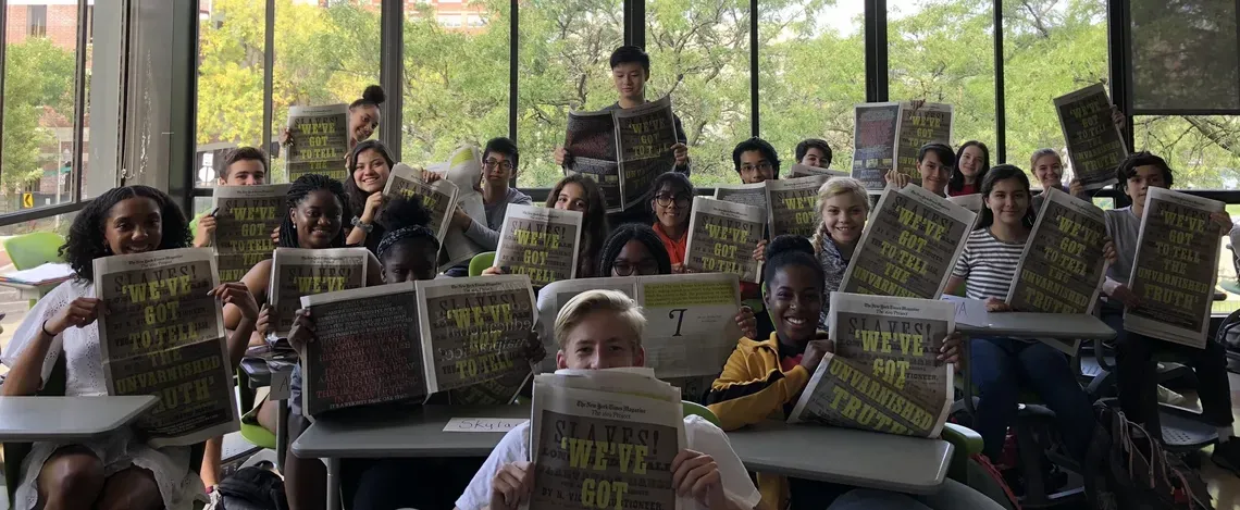 a group of students in a classroom hold up their paper copies of The 1619 Project
