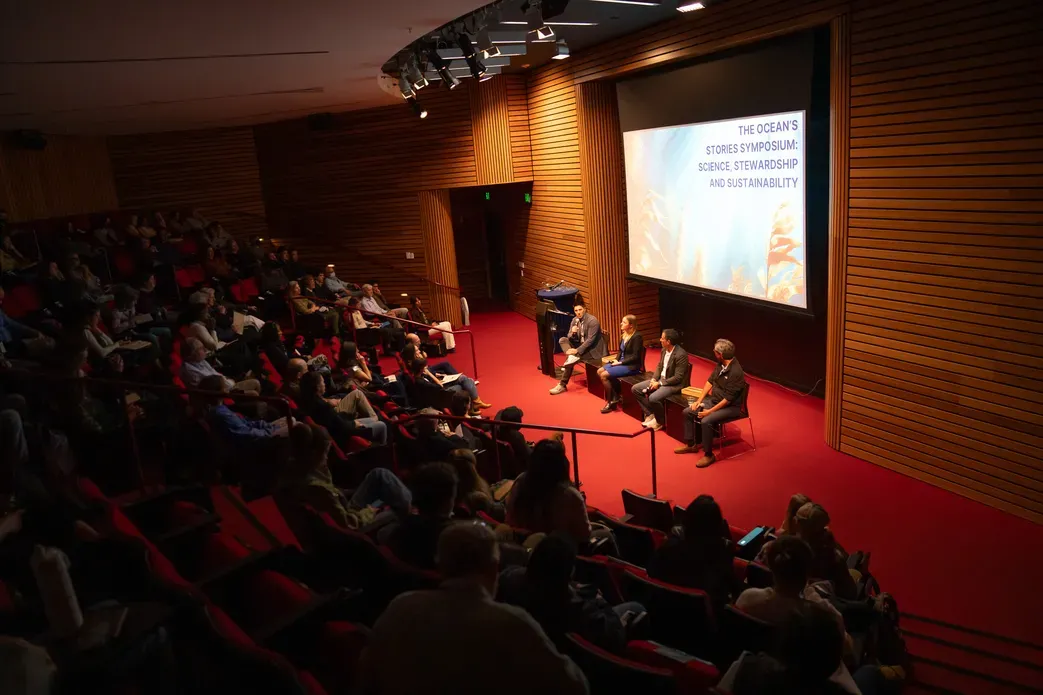 Four man sit for a panel in front of screen in auditorium