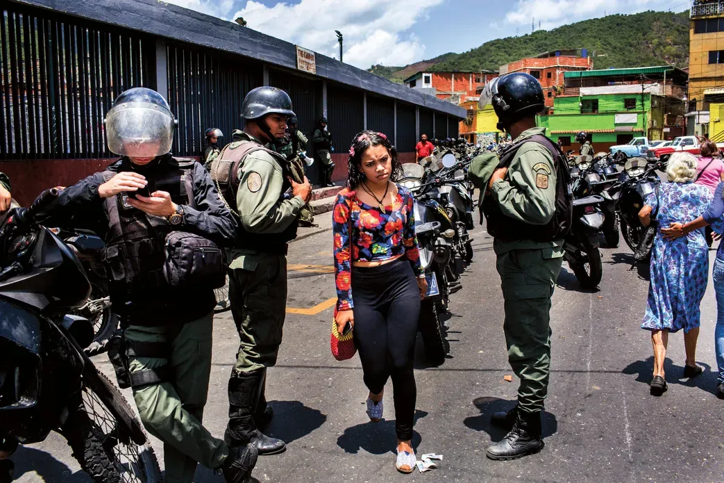 A young woman looks directly at the camera as police in military gear surround her, with colorful buildings in the background