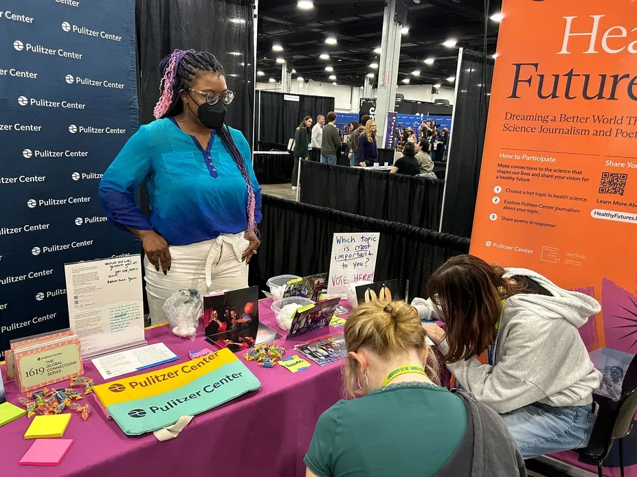 A person stands behind a table at an exhibit booth, talking to students who are seated and writing.