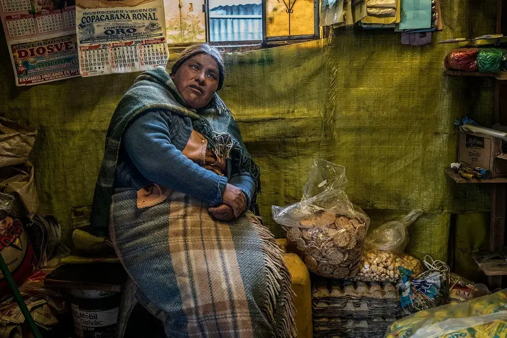A resident, bundled up against the constant Andean cold, sits in her food stall in the gold-processing area. This part of La Riconada is most acutely contaminated with mercury, which condenses on everything after being vaporized during the gold-purification process. Peru, 2019. Image by James Whitlow Delano.