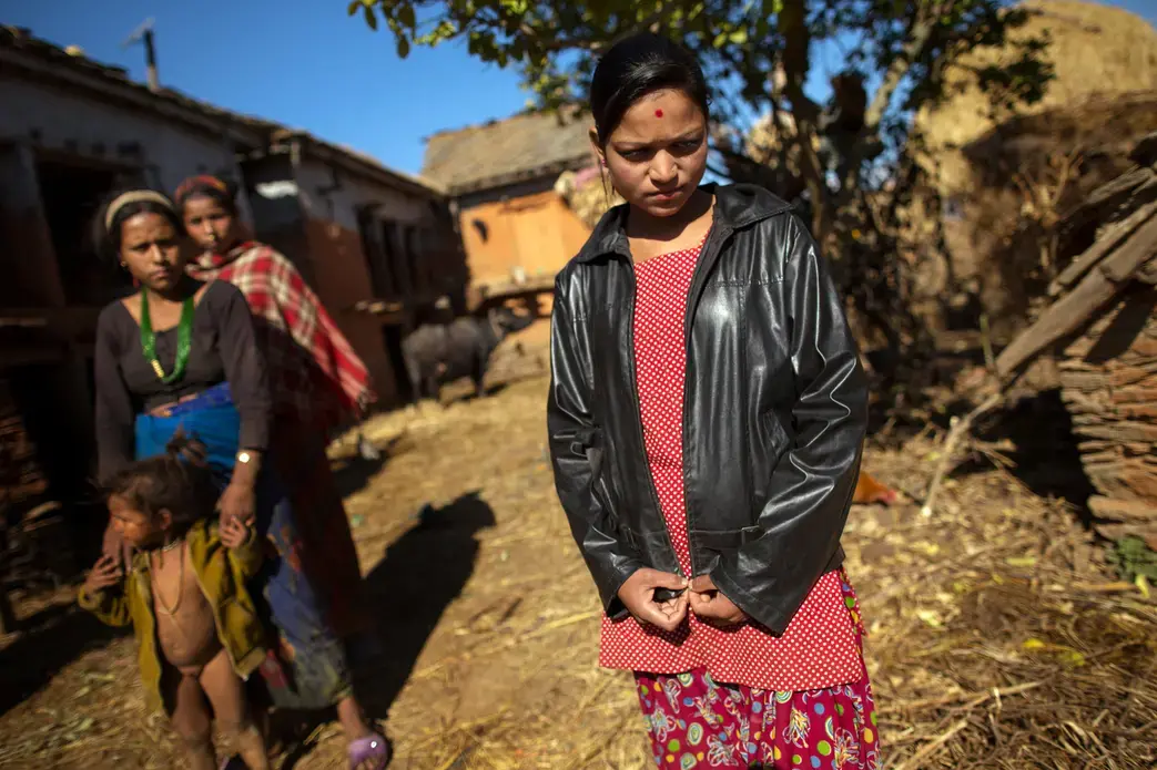 Rabina Kumari Nepali, 14, stands apart from her family in front of the chaupadi goth (right), a squat hut where she sleeps during her period. Women in this region are considered unclean while they are menstruating and must live apart from their families during those days. They find shelter in similar huts, caves or out in the open. Image by Allison Shelley. Nepal, 2012. Media file: MK3_8686web.jpg