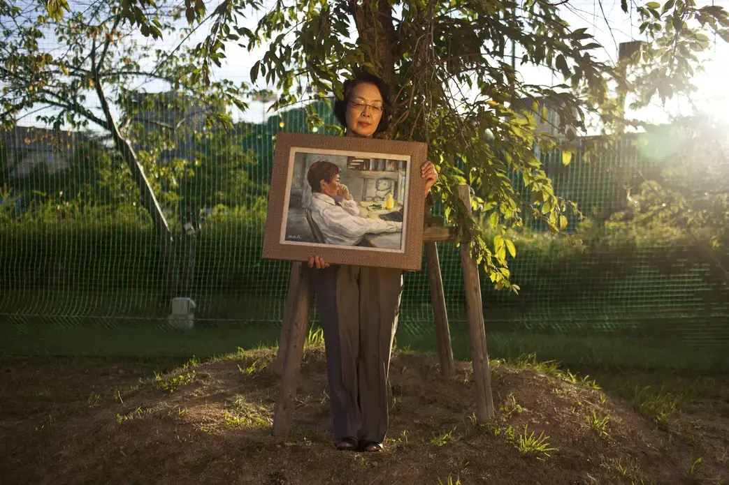 Michiyo Nishigaki, 67, with a painting of her son, poses under the cherry tree she planted after his death. Her son Naoya, a systems engineer who suffered from depression brought on by excessive overwork, killed himself in 2006. Image by Shiho Fukada. Japan, 2012. Media file: Nishigaki137.jpg
