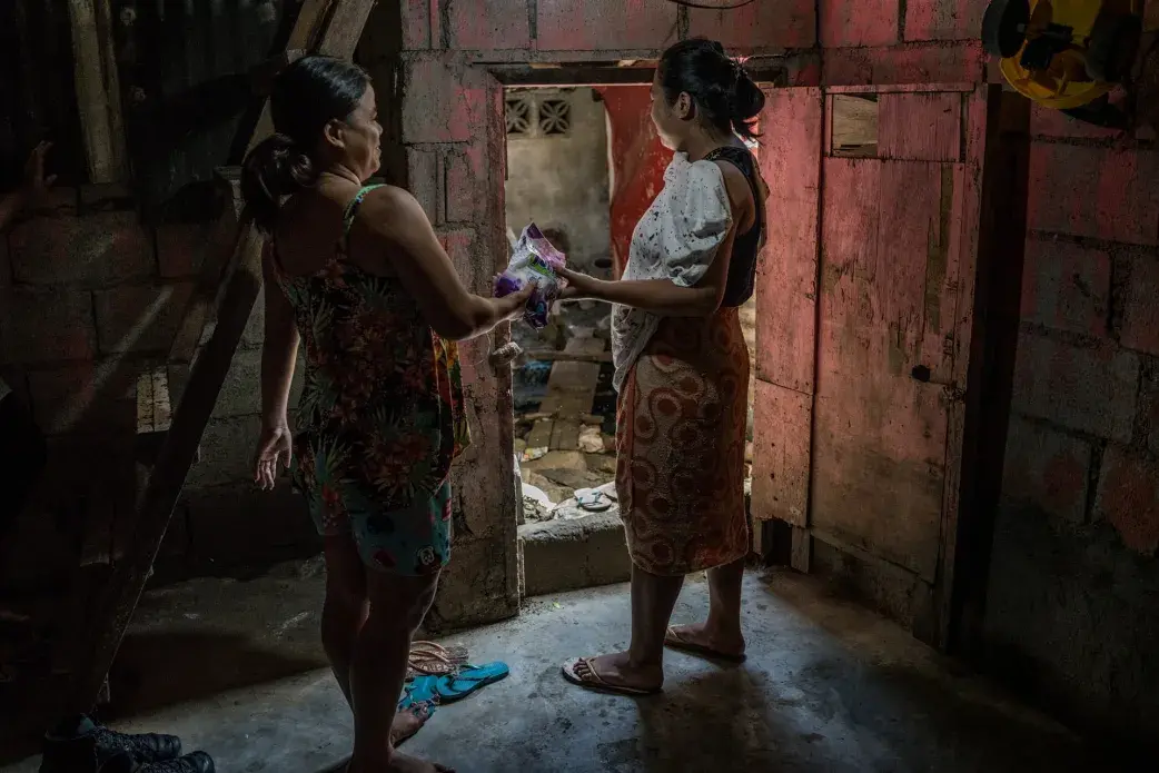 Rhoda hands over an Avon purchase to her neighbour, as her business has begun to slowly expand, on the first floor of her house, she was forced to abandon due to recurring flooding. Image by James Whitlow Delano. Philippines, 2018. 