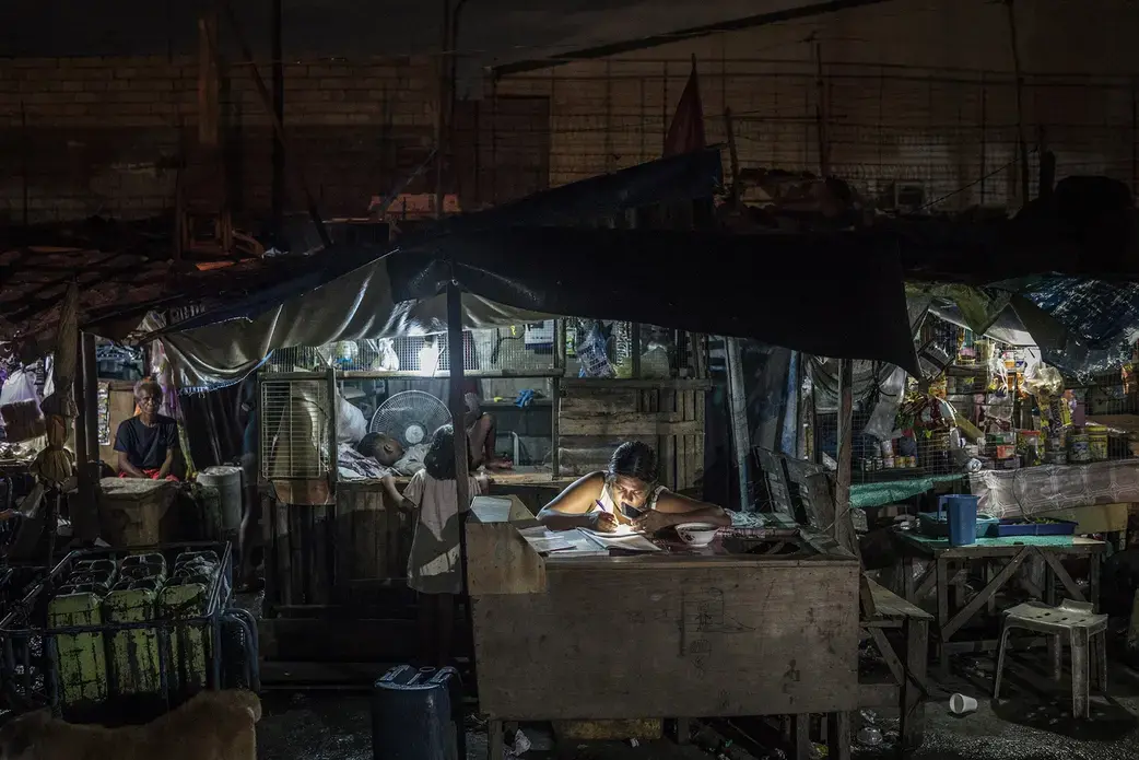 A woman works on bookkeeping using the light of her phone in the Market 3 slum in the Navotas Fish Port Complex. Market 3 has electricity but no plumbing. Residents often turn to drugs to alleviate hunger and stay awake for the long hours they work at low pay. Image by James Whitlow Delano. Philippines, 2017. A woman works on bookkeeping using the light of her phone in the Market 3 slum in the Navotas Fish Port Complex. Market 3 has electricity but no plumbing. Residents often turn to drugs to alleviate hunger and stay awake for the long hours they work at low pay. Image by James Whitlow Delano. Philippines, 2017.