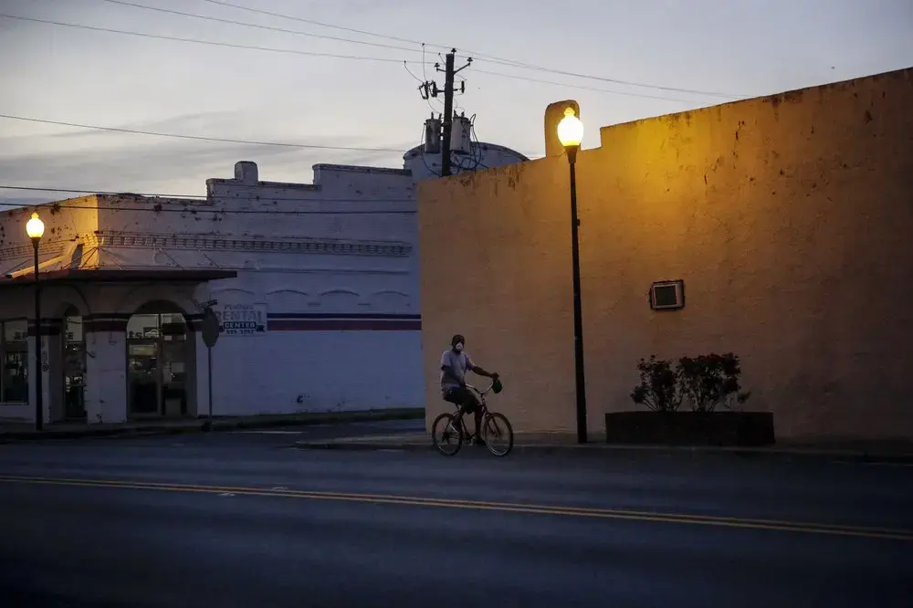 A man riding a bicycle down an empty street wears a protective mask amid the COVID-19 coronavirus outbreak on Friday, April 17, 2020, in Dawson, Ga. Image by Brynn Anderson / AP Photo. United States, 2020.