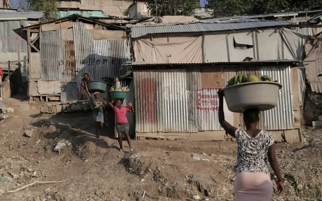 Women carrying produce and other goods walk by some of the tin shacks that make up the Teren Toto camp in Haiti’s capital. Image by Jose A. Iglesias. Haiti, 2019.