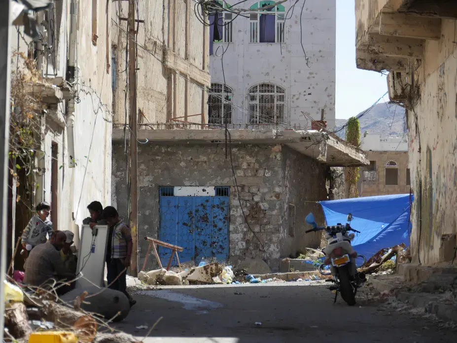 Children gather around their father as he fixes a solar panel for electricity. A blue tarpaulin in the background blocks the line of sight of pro-Houthi snipers in the no-mans-land between the warring factions who have been battling for control of the city of Taiz since May 2015. Image by Iona Craig. Yemen, 2017.