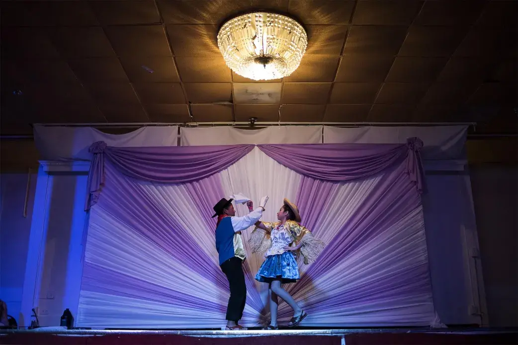 A young boy and girl represent their dance group, Tradiciones Bolivianas (Bolivian Traditions), dancing a traditional cueca at a children's talent show, Miss and Mister Talent, in Fairfax, Virginia. The talent competition includes young boys and girls from different south and Central American countries, but only Bolivia has folkloric contestants and dance groups. More than five children’s dance groups perform at the event. Many Bolivian parents share stories of their children dancing at 3 or 4 years old. They say that dancing “is in their blood.” Image by Carey Averbook. United States, 2016.