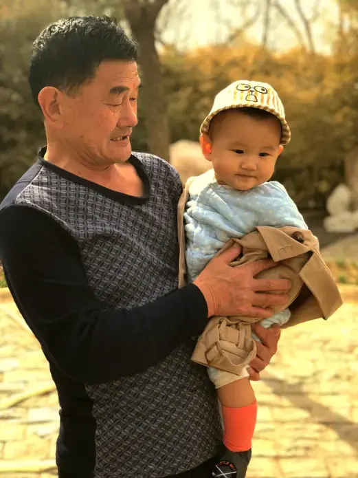 At a park in Beijing, an elderly woman rolls a stroller while this man carries a baby boy. Image by Argentina Maria-Vanderhorst. China, 2018.