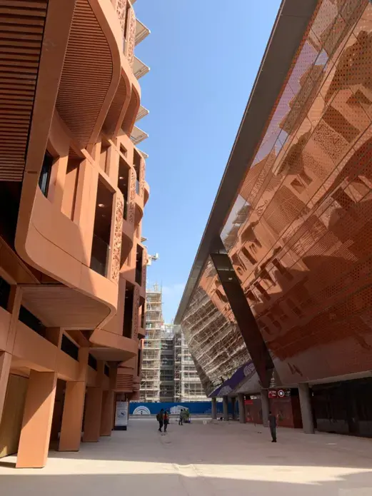 Masdar City’s streets are narrow and lined with buildings that promote cool street temperatures. The terracotta residential building on the left has screens which shade the rooms inside. The incubator building to the right slopes outward to further shade the streets below. Construction continues in the background. Image by Anna Gleason. United Arab Emirates, 2019.