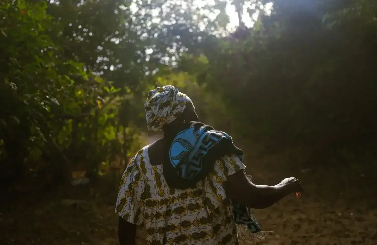 A woman faces away from the camera and walks into the sun shining down on a forest with her arms raised